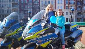 Na zdjęciu na policyjnym motocyklu siedzi dziewczynka, obok stoi kobieta.